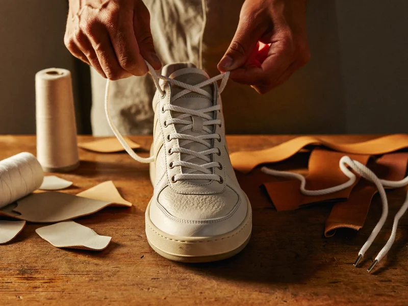 Hände schnüren einen Sneaker an einer Werkbank, daneben Lederreste und Gummisohlen.
